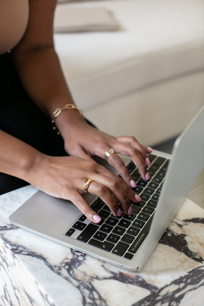 Close-up of a woman’s hands typing on a silver laptop, styled with gold rings and soft pink nails on a marble desk.